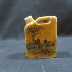 Può includere: Fiaschetta in ceramica color senape con manico e beccuccio bianco. La fiaschetta presenta una scena dipinta di persone a cavallo con cani e un albero. La scena è ambientata in un campo con una tavolozza di colori marrone e verde.