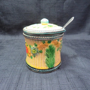 May include: A vintage ceramic jar with a lid and spoon. The jar is yellow with vertical stripes and floral designs in orange, blue, and green. The lid is light yellow with a blue trim and a small blue knob. A silver spoon rests on the lid.