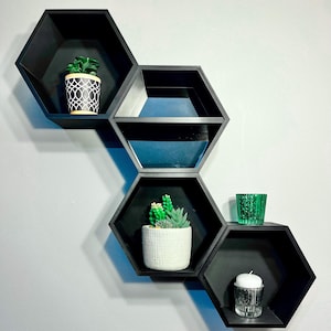 Könnte beinhalten: Schwarze sechseckige Wandregale mit dekorativen Gegenständen. Die Regale beherbergen eine Topfpflanze mit einem schwarz-weißen Design, einen kleinen Kaktus in einem weißen Topf, ein grünes Glas und eine Kerze in einem Glashalter.