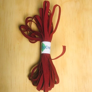 Könnte beinhalten: Eine Rolle maroonfarbenes Gummiband, etwa 1,2 cm breit, ist auf einer Holzoberfläche aufgerollt. Das Gummiband ist mit "Big Craft" beschriftet.