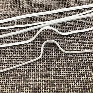 May include: A close-up of several white plastic nose wire pieces for face masks. The nose wires are bent into a U-shape and are lying on a textured gray surface.