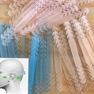 May include: A close-up image of a large quantity of clear plastic ear savers for face masks. The ear savers are in two colors: light blue and clear. The ear savers are arranged in a random pattern. The text "Ship Within 1 day" is visible in the upper right corner of the image.