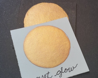 May include: A close-up of two circular swatches of metallic gold paint on a black surface. The bottom swatch is on a light gray card with the handwritten text "sunset glow". The top swatch is partially visible.