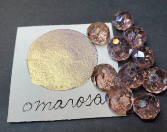 May include: A close-up of a gold metallic paint sample with the word "omarosal" written in black ink. The sample is surrounded by iridescent purple glass beads.