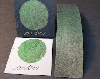 May include: Two swatches of green shimmery paint, one on a black card and one on a white card. Both swatches are labeled "arden".