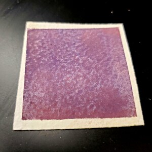 May include: A square watercolor painting with a textured, purple and pink gradient. The artwork is bordered by a white, textured paper edge. The painting is on a black surface.
