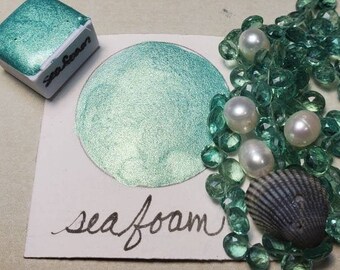May include: A teal watercolor paint swatch with the handwritten text "sea foam" next to a small paint block labeled "Sea Foam". The image includes a seashell, pearls, and green glass beads.