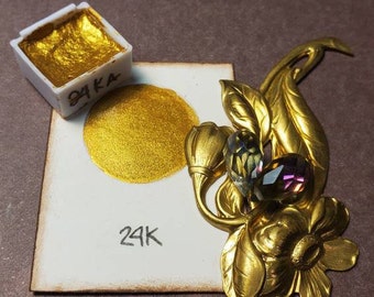 May include: A close-up of a gold-colored watercolor paint swatch and a decorative gold-toned floral brooch with two iridescent gemstones. The paint is labeled "24K" on the swatch and the paint container.
