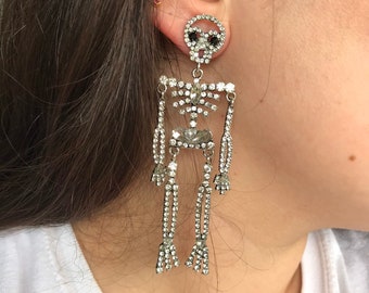 May include: A pair of silver skeleton earrings with crystal accents. The earrings are long and dangle, with a skull at the top and a skeleton body below.