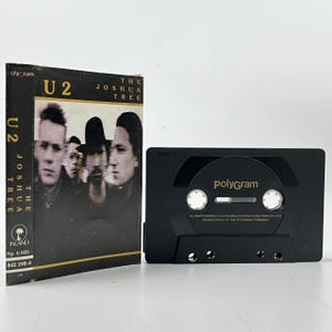 May include: A black cassette tape with the text "PolyGram" printed on the front. The cassette tape is in a plastic case with a black and white image of the band U2 on the front. The text "U2 The Joshua Tree" is printed on the case.