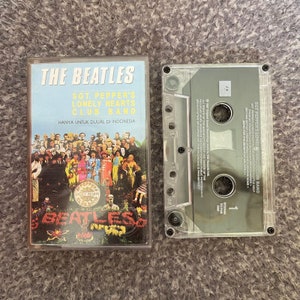 Könnte beinhalten: Eine Vintage-Kassette des Beatles-Albums "Sgt. Pepper's Lonely Hearts Club Band". Das Albumcover zeigt eine farbenfrohe Gruppe von Menschen. Die Kassette befindet sich in einer transparenten Kunststoffhülle mit Text in Englisch und Indonesisch.
