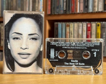May include: A cassette tape of Sade's "The Best Of Sade" album. The cassette case features a black and white portrait of Sade. The album's tracklist is visible on the cassette label. The cassette is clear.