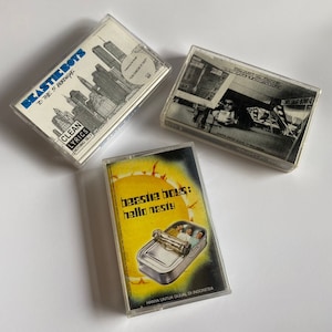 Puede incluir: Tres cintas de cassette con diferentes portadas de álbum. La primera cinta es un álbum de Beastie Boys titulado "To the 5 Boroughs" con un fondo de paisaje urbano. La segunda cinta es un álbum de Beastie Boys titulado "The Sounds of Science" con una foto de un hombre en un coche. La tercera cinta es un álbum de Beastie Boys titulado "Hello Nasty" con un sol amarillo y una lata de sardinas con los miembros de la banda dentro.