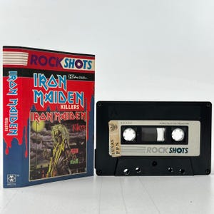 Puede incluir: Una cinta de casete negra con una etiqueta blanca. La etiqueta tiene el texto "Rock Shots" y "A HMG Collection Production". La cinta de casete es una grabación musical del álbum "Killers" de la banda Iron Maiden.