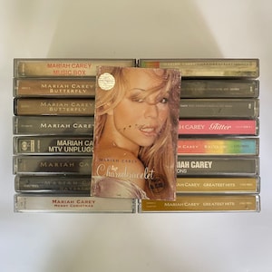 Może przedstawiać: Stos kaset magnetofonowych Mariah Carey, w tym tytuły takie jak "Music Box", "Butterfly", "Charmbracelet", "Glitter" i "Greatest Hits".