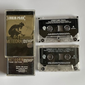 以下が含まれることがあります： リンキン・パークのアルバム「Meteora」の3つのカセットテープ。最初のテープは標準版、2番目はツアー版、3番目はボーナスライブトラック付きのツアー版です。テープはすべて透明なプラスチック製で、黒いラベルとテキストが書かれています。