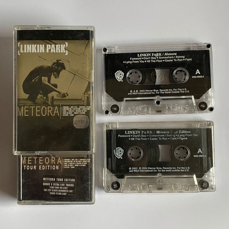 以下が含まれることがあります： リンキン・パークのアルバム「Meteora」の3つのカセットテープ。最初のテープは標準版、2番目はツアー版、3番目はボーナスライブトラック付きのツアー版です。テープはすべて透明なプラスチック製で、黒いラベルとテキストが書かれています。