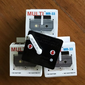 以下が含まれることがあります： 黒い文字で書かれた白いカセットテープケースが3つ。ケースには「MULTI MR-33」と表示されています。白いレバーと赤い矢印が付いた黒いプラスチック製の装置がケースの上に置かれています。