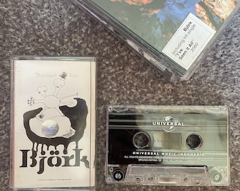 Op de afbeelding: Drie cassettebandjes, waarvan twee van doorzichtig plastic met zwarte labels zijn en één wit is met een zwart-witte illustratie van een wezen met drie hoofden. De witte cassetteband heeft de tekst "Greatest Hits" en "Björk" op het label. Het zwarte label op de doorzichtige cassetteband zegt "Universal Music Indonesia". De andere doorzichtige cassetteband heeft de tekst "Björk" en "Including hit single "I've Seen It All" 2060" op het label.