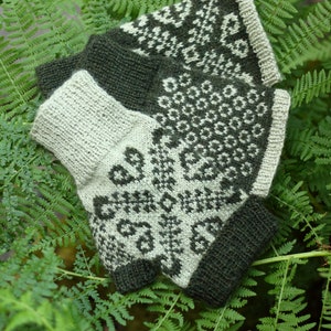 May include: A pair of knitted fingerless gloves with a geometric pattern in shades of green and white. The gloves are laid out on a bed of green ferns.