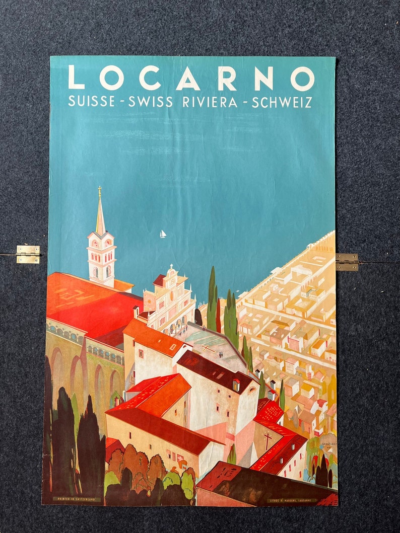 May include: Vintage travel poster advertising the Swiss Riviera town of Locarno. The poster features a colorful illustration of the town's architecture, including a church with a tall spire, red-roofed buildings, and palm trees. The text "Locarno" is in large white letters at the top of the poster, and the text "Suisse - Swiss Riviera - Schweiz" is in smaller white letters below it.