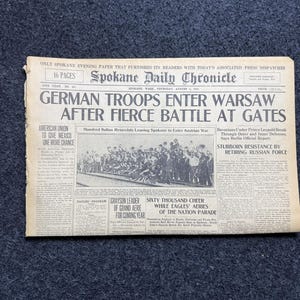 Puede incluir: Un periódico antiguo, el Spokane Daily Chronicle, con titulares sobre la entrada de tropas alemanas en Varsovia después de una batalla. El periódico está envejecido, con texto visible y una fotografía en blanco y negro. El titular dice "GERMAN TROOPS ENTER WARSAW AFTER FIERCE BATTLE AT GATES."