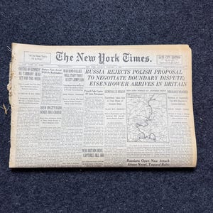 Puede incluir: Un ejemplar antiguo del periódico The New York Times, con titulares sobre Rusia, Polonia y Eisenhower. El periódico está envejecido y desgastado, sobre una superficie azul oscuro.