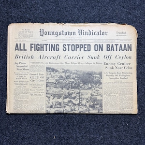 May include: An aged copy of the Youngstown Vindicator newspaper, with headlines about the end of fighting on Bataan and naval battles. The paper is yellowed with age and lies on a dark blue surface.