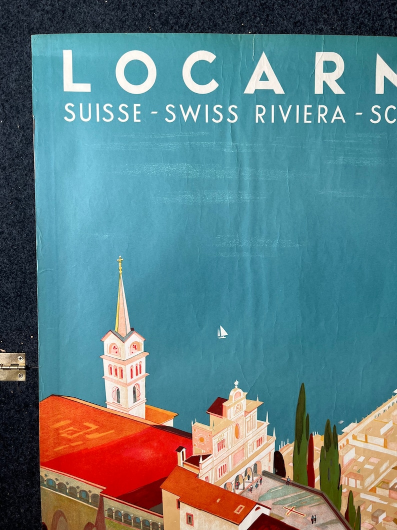 May include: A vintage travel poster advertising the Swiss Riviera town of Locarno. The poster features a colorful illustration of the town's architecture, including a church with a tall spire and a view of the lake. The text on the poster reads "Locarno Suisse - Swiss Riviera - Schweiz - Svizzera - Svizzera Italiana".