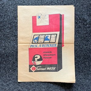 May include: A red, white and blue mock election issue of Junior Current Week magazine. The cover features a slot machine with three options: a fish, a man with glasses, and a donkey. The slot machine has a question mark on the side and the text "Pick a Winner" below it.