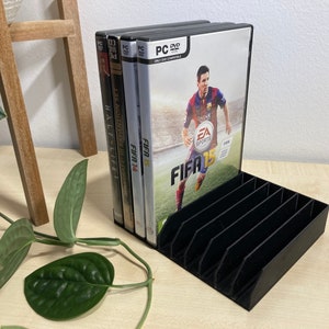 Può includere: Un organizer per DVD in plastica nera che contiene diverse custodie di videogiochi, tra cui "FIFA 15" e "Half-Life 2". L'organizer ha più slot per l'archiviazione ed è progettato per mantenere DVD e videogiochi organizzati. Le custodie sono compatibili con PC DVD.