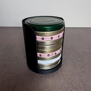 Puede incluir: Un conjunto de latas redondas de metal con bandas rosas y blancas, sujetas en un soporte negro. La lata superior es verde con texto dorado. Las bandas tienen un diseño de flechas repetidas. Las latas están apiladas.
