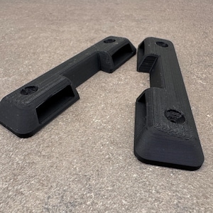 May include: Two black, rectangular 3D-printed objects. Each has a rectangular cutout and a circular hole. The objects are on a neutral-toned surface. The items appear to be components or parts.