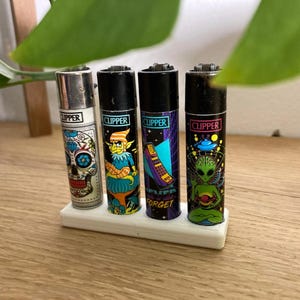 May include: Four different Clipper lighters with colorful designs. The lighters are in a white plastic holder. The designs include a skull, a goblin, a spaceship, and an alien.