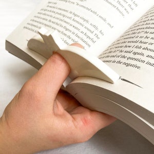 Puede incluir: Un sujeta páginas de plástico blanco en forma de pájaro con las alas extendidas. El sujeta páginas se utiliza para mantener un libro abierto.