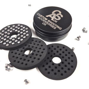 May include: Black circular shooting accessories with a central hole and numerous smaller holes. A black tin with the text "AS UK CUSTOM SHOOTING ACCESSORIES" is also present, along with scattered pellets. The accessories appear to be for air guns.