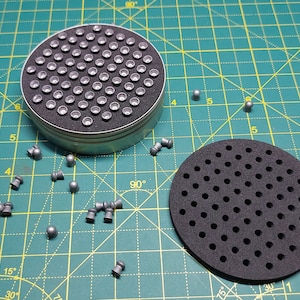 May include: A round metal container filled with small silver pellets, a black foam pad with holes, and several loose silver pellets on a green grid surface.