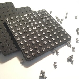 May include: Four black foam squares with circular indentations, each filled with a silver-colored pellet. Scattered silver-colored pellets on a white surface.