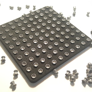 May include: A black square-shaped pellet tray with a grid of round indentations. The tray is surrounded by a scattering of silver-colored round pellets.