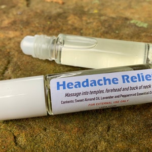 May include: Two clear glass roll-on bottles with white caps. One bottle has a label that reads "Headache Relief" with instructions and ingredients. The bottles are filled with a clear liquid and sit on a textured, brown surface.
