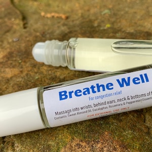 May include: A white roller bottle with a blue label that reads "Breathe Well" for congestion relief. The label also lists the ingredients: Sweet Almond Oil, Eucalyptus, Rosemary & Peppermint Essential Oils. The bottle is on a brown and green textured surface.
