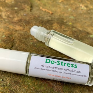 May include: Two white roller bottles with clear liquid inside. The label on the front bottle reads "De-Stress" and includes instructions to "Massage into temples and back of neck". The label also lists the ingredients: "Sweet Almond Oil, Clary Sage, Lavender & Lemon Essential Oils". The label also states "FOR EXTERNAL USE ONLY" and includes the name "LINDA ODOM".