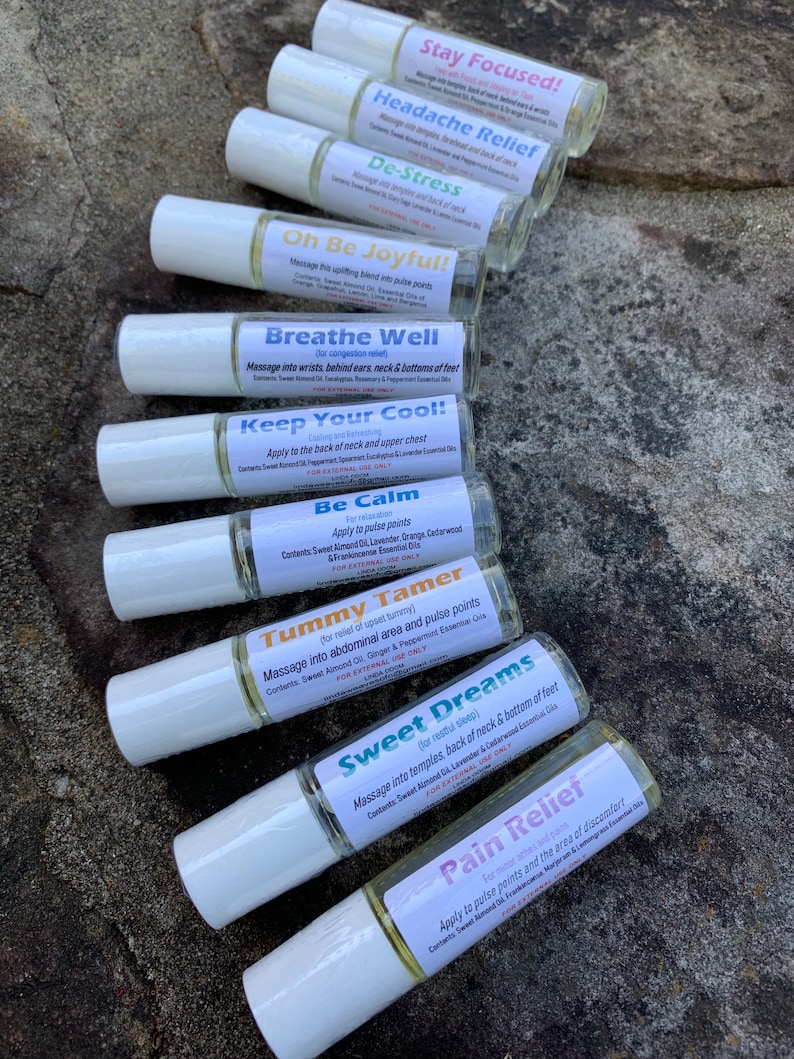 May include: A collection of roll-on essential oil blends with white caps and labels. The labels feature text such as "Stay Focused!", "Headache Relief", and "De-Stress". The bottles are arranged on a textured, natural surface.