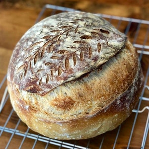 May include: A freshly baked loaf of sourdough bread with a decorative leaf pattern on the top crust. The bread is golden brown and has a rustic appearance.