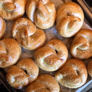 May include: A baking sheet filled with freshly baked soft pretzels. The pretzels are golden brown and have a shiny glaze. Some of the pretzels have coarse salt sprinkled on top.