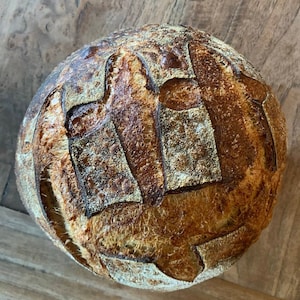May include: A close-up of a round loaf of sourdough bread with a deep brown crust and a pattern of slashes on the top.