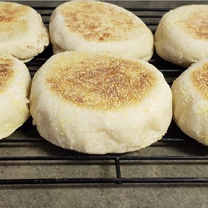 May include: Six freshly baked English muffins on a wire rack. The muffins are golden brown and have a slightly crispy exterior.