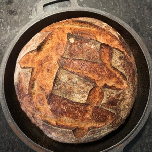 May include: A freshly baked loaf of sourdough bread with a crusty exterior and a soft, airy interior. The bread is sitting in a black cast iron pan.