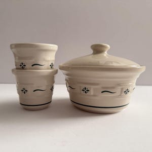 May include: A set of cream-colored ceramic kitchenware. The set includes two small, stacked pots and a larger lidded casserole dish. Each piece is adorned with a dark green floral pattern and a curved line. The pieces are arranged on a white surface.