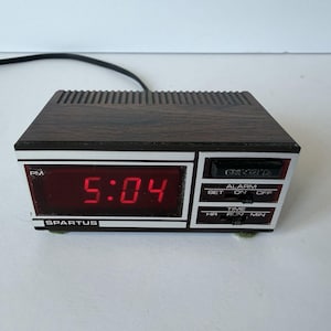 May include: A vintage digital alarm clock with a dark wood-grain top and a red LED display showing 5:04 PM. The clock face is framed in white and silver, with the brand name "SPARTUS" visible. It has alarm and time setting controls.